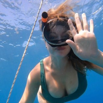 Woman in green swimsuit snorkeling underwater, waving with one hand near a rope.